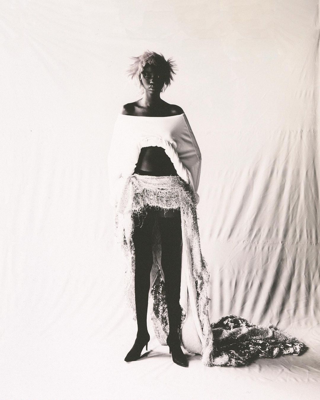 Monochrome portrait of a model with short textured hair wearing an off-the-shoulder white top and a long sheer overlay skirt