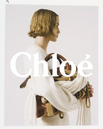 Fashion portrait of a woman with short blond hair, wearing an off-white knit sweater and holding a tan leather bag in a studio setting.