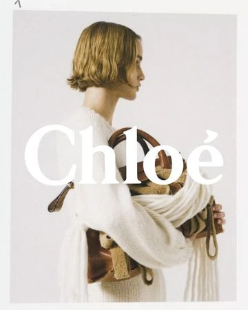 Fashion portrait of a woman with short blond hair, wearing an off-white knit sweater and holding a tan leather bag in a studio setting.