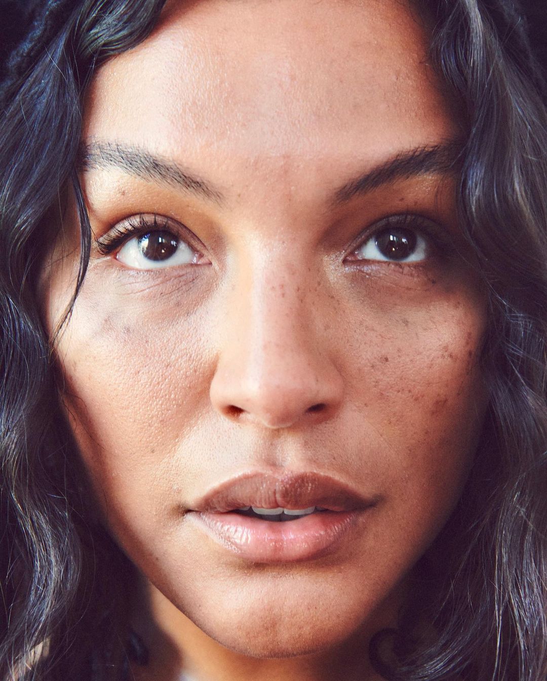 Close-up portrait of a woman with dark, wavy hair and brown eyes looking upward with a neutral expression. Skin is fair with visible freckles.