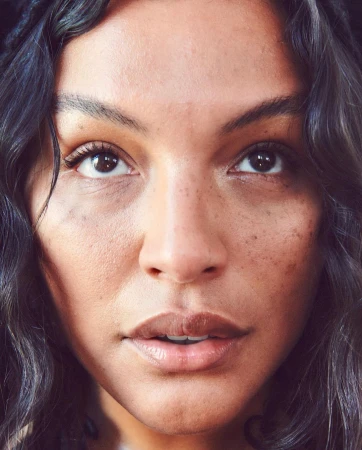 Close-up portrait of a woman with dark, wavy hair and brown eyes looking upward with a neutral expression. Skin is fair with visible freckles.