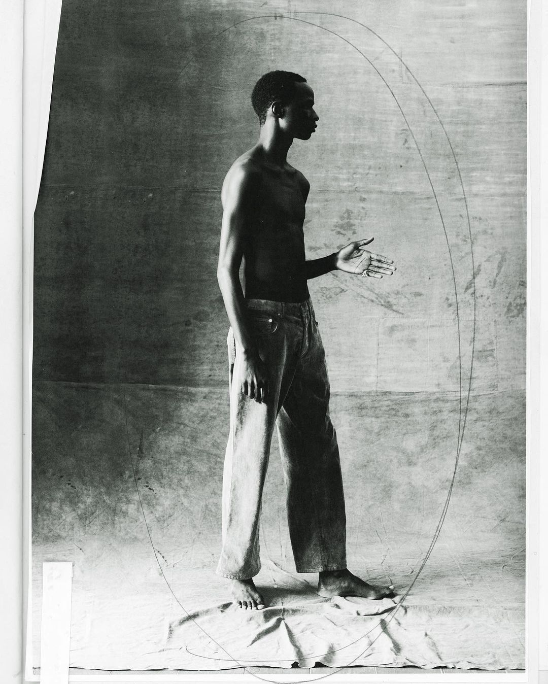 Monochrome studio shot of a bare-chested adult male in jeans standing with hand extended against a neutral background with drawn lines.