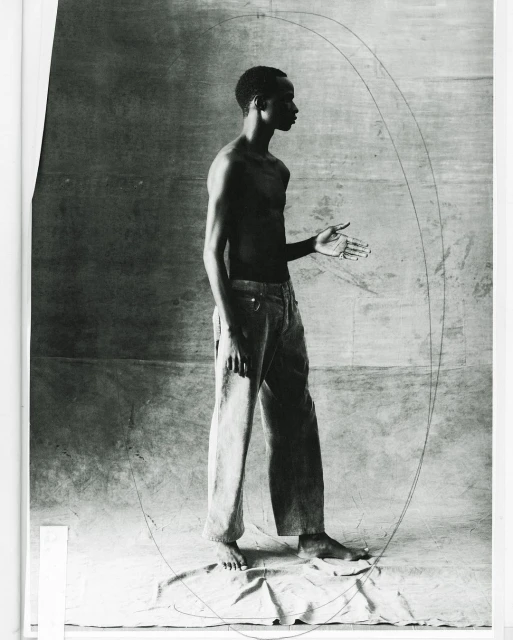 Monochrome studio shot of a bare-chested adult male in jeans standing with hand extended against a neutral background with drawn lines.
