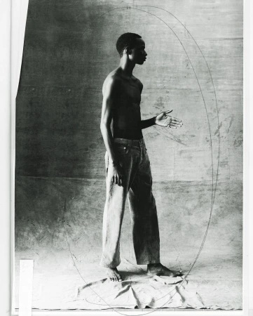 Monochrome studio shot of a bare-chested adult male in jeans standing with hand extended against a neutral background with drawn lines.
