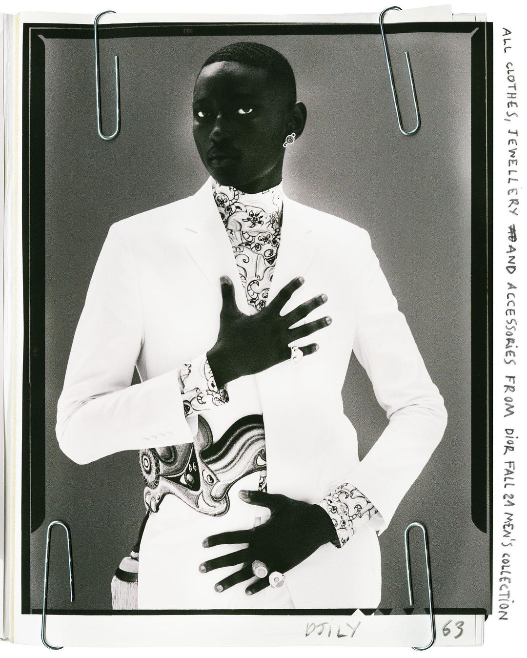 Fashion shot of a Black male model wearing a white suit with patterned shirt and jewellery, posing in front of a gray backdrop.