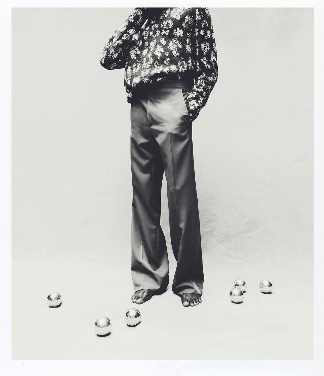 Monochrome studio shot of a stylish adult standing barefoot among scattered metal spheres in an artistic fashion photograph.