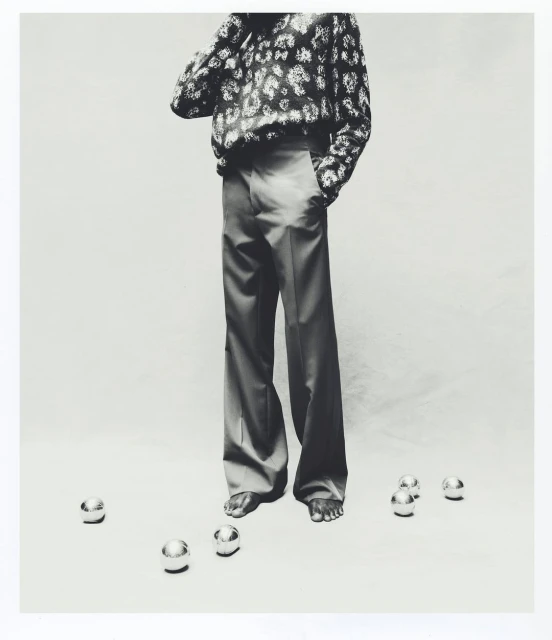 Monochrome studio shot of a stylish adult standing barefoot among scattered metal spheres in an artistic fashion photograph.