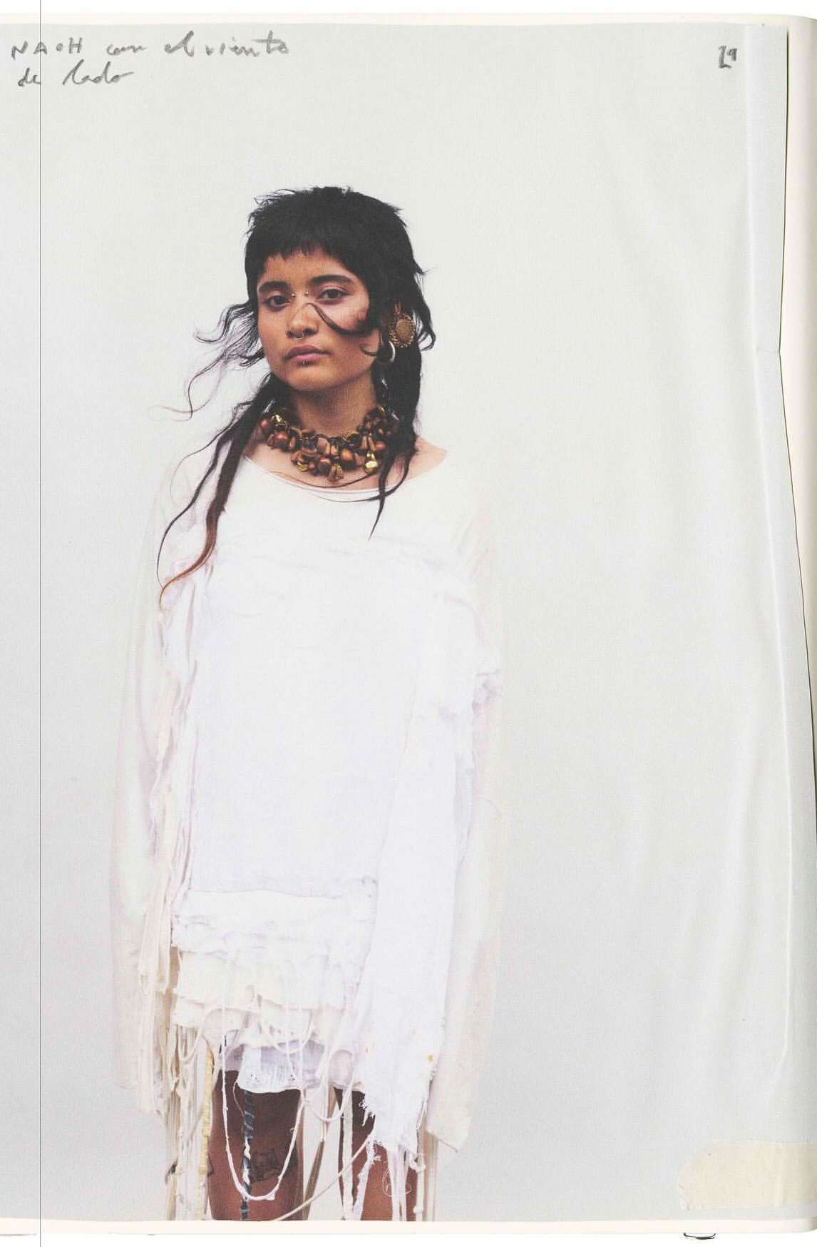 Portrait of a person with dark hair and a fringe, wearing a white dress and beaded necklace against a plain studio backdrop.