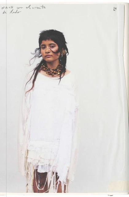 Portrait of a person with dark hair and a fringe, wearing a white dress and beaded necklace against a plain studio backdrop.