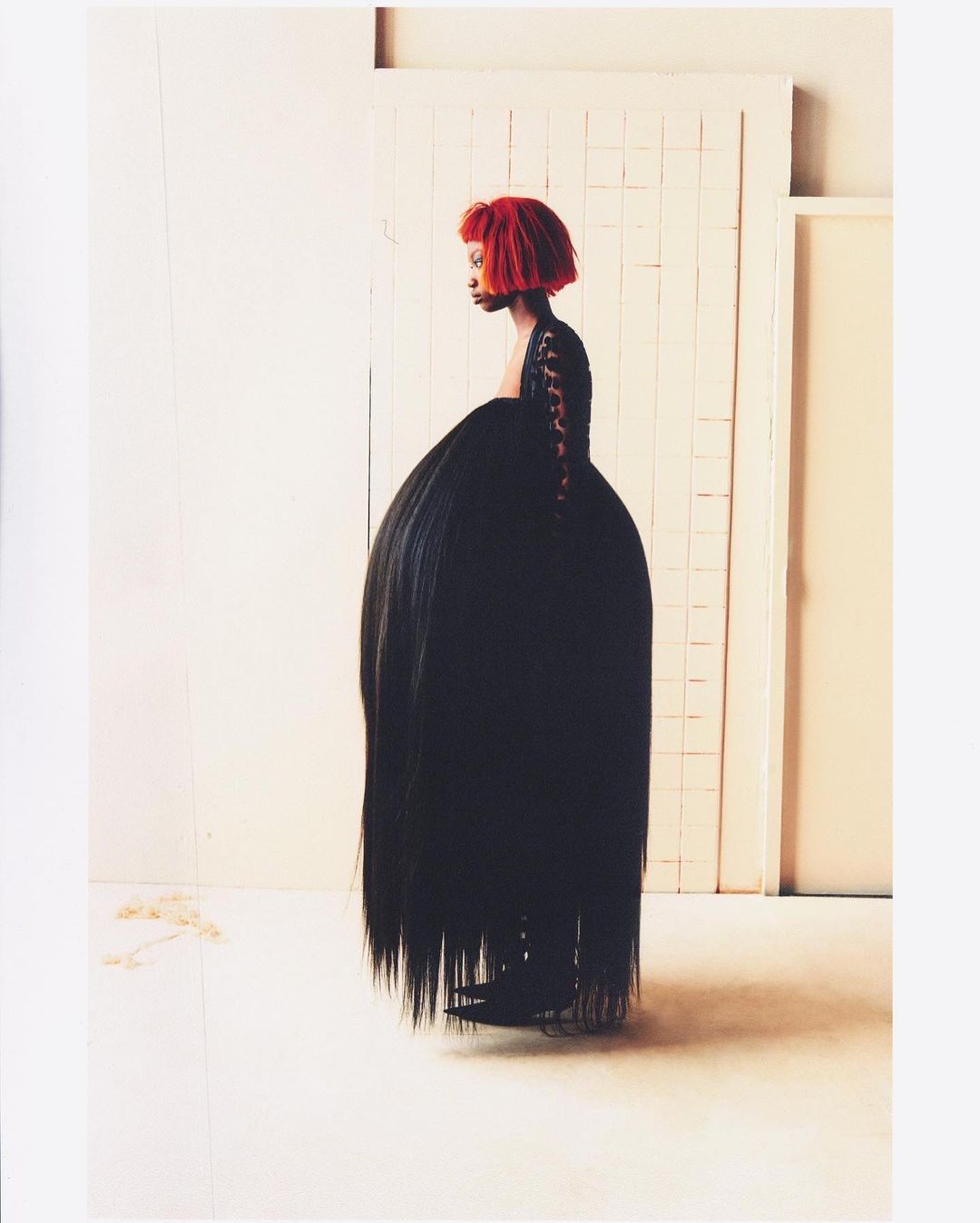 A woman with a red hairstyle poses in a dramatic black dress with a voluminous skirt in a minimalist studio setting, fashion forward and unique.