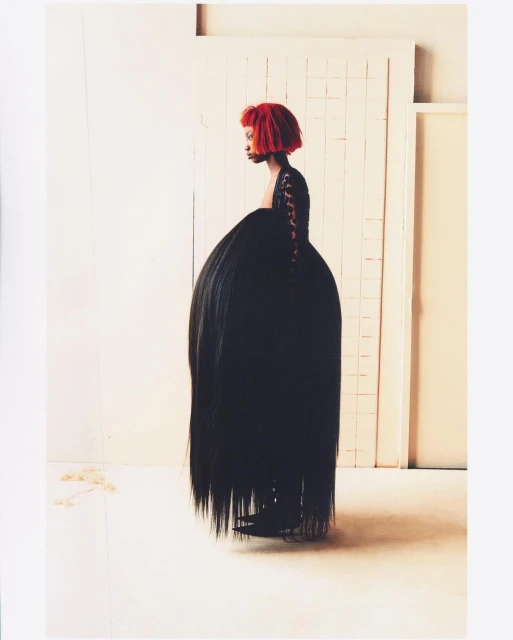 A woman with a red hairstyle poses in a dramatic black dress with a voluminous skirt in a minimalist studio setting, fashion forward and unique.