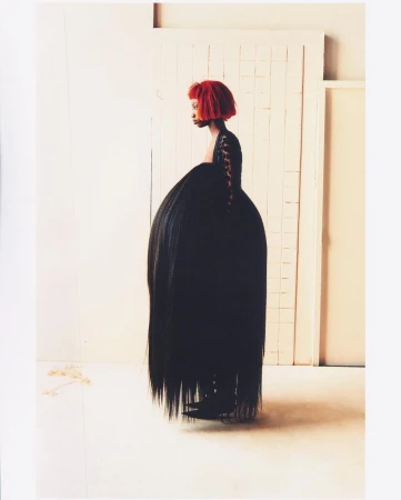 A woman with a red hairstyle poses in a dramatic black dress with a voluminous skirt in a minimalist studio setting, fashion forward and unique.