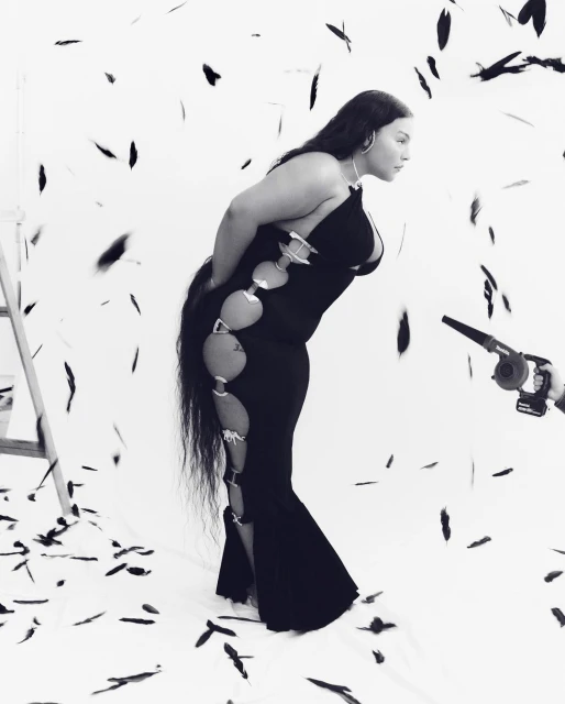 A woman in a black dress poses in a studio filled with black feathers, with a blower held by a person on the side.