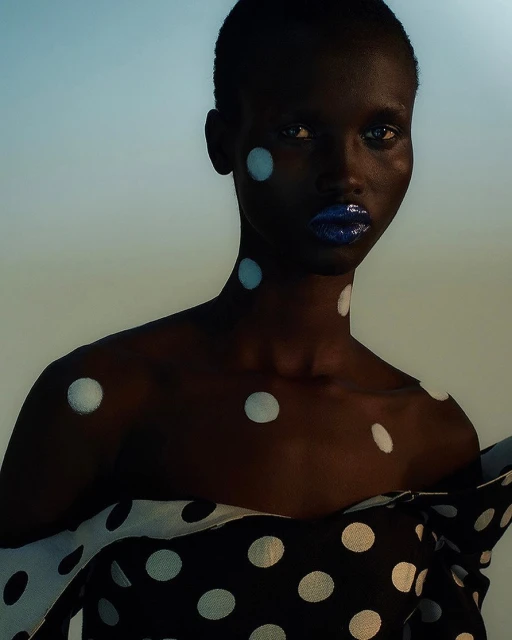 Portrait of a dark-skinned woman with blue lipstick and polka dot body art and clothing, set against a gradient blue and beige background.