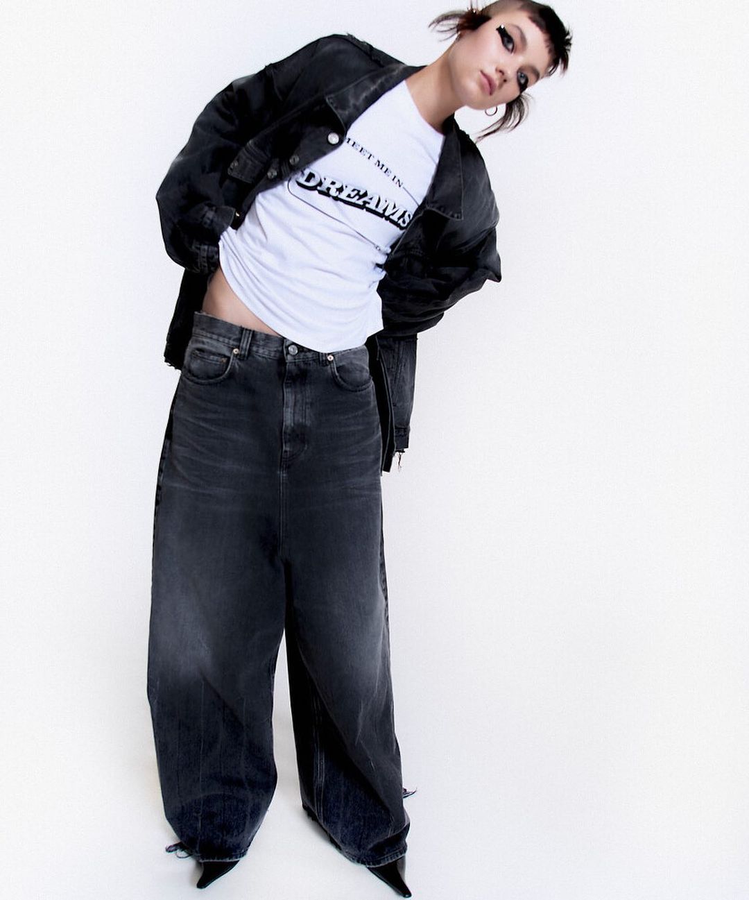 Young adult woman wearing oversized denim jacket and jeans, white t-shirt, and black heels posing in a bright studio setting.