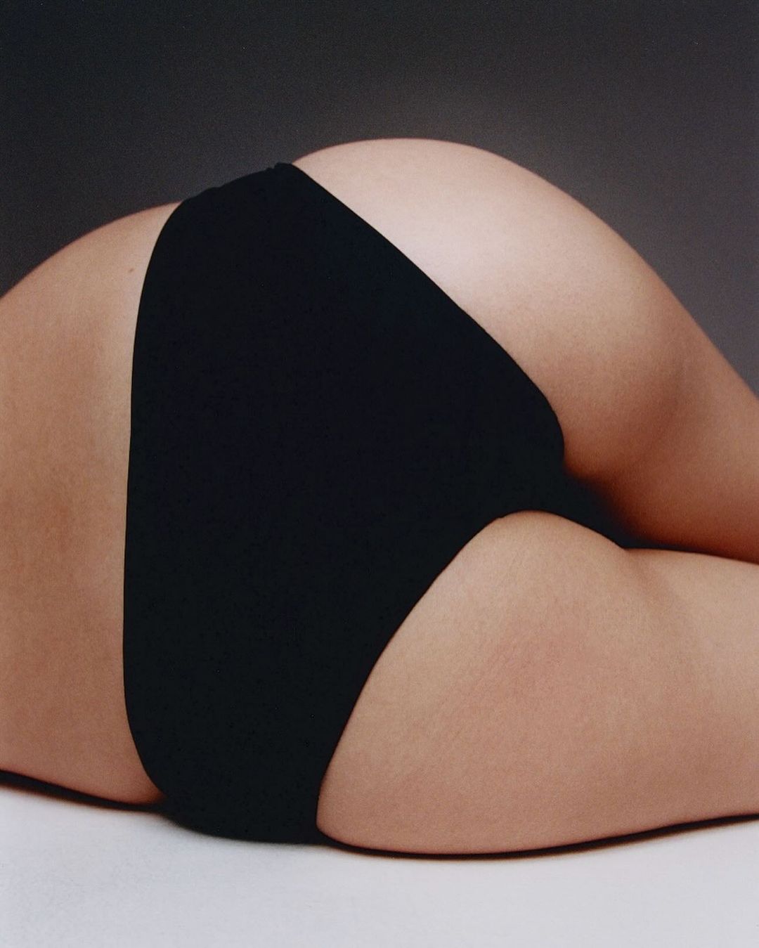 Close-up view of a person lying down, featuring black underwear and skin details against a dark gray studio background.