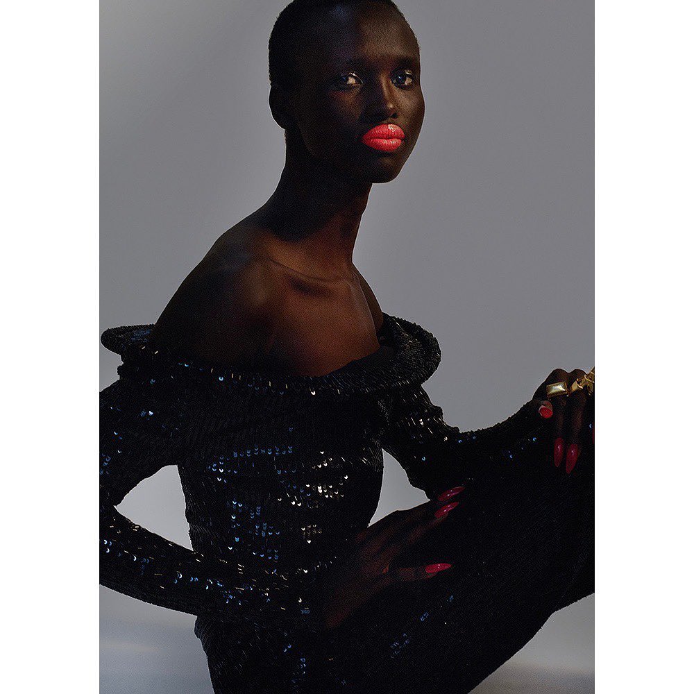 Glamorous portrait of a dark-skinned woman in a sequined dress with coral lipstick, posed against a solid light gray backdrop.