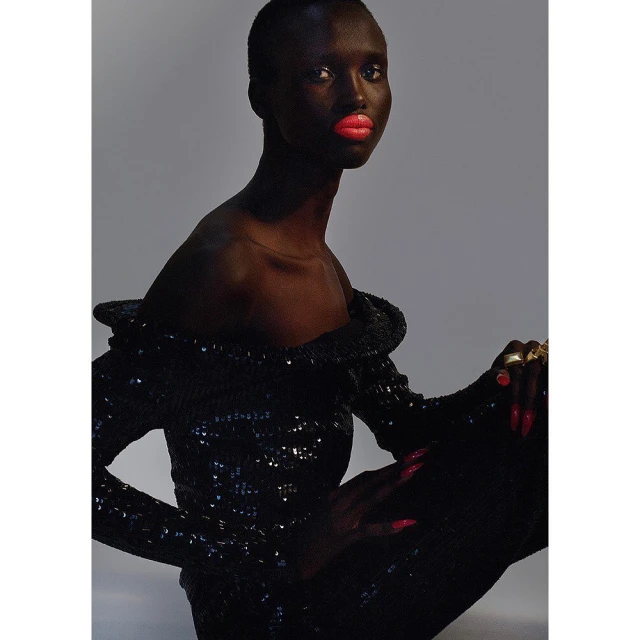 Glamorous portrait of a dark-skinned woman in a sequined dress with coral lipstick, posed against a solid light gray backdrop.