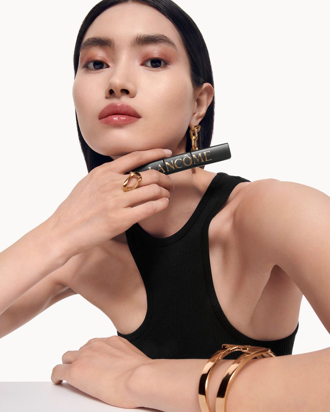 Close-up portrait of an East Asian woman wearing a black tank top, gold jewelry, and holding a Lancome lipstick tube.