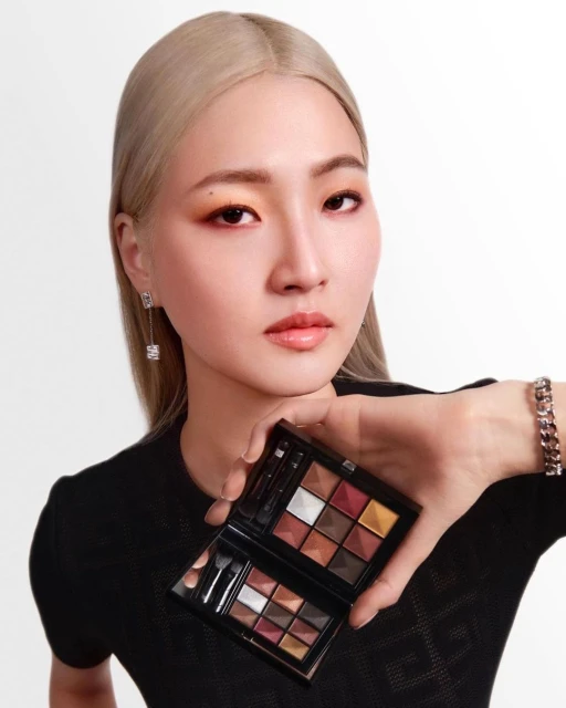 A woman with fair skin and light blonde hair showcases a makeup palette with various shades in a beauty or fashion context.