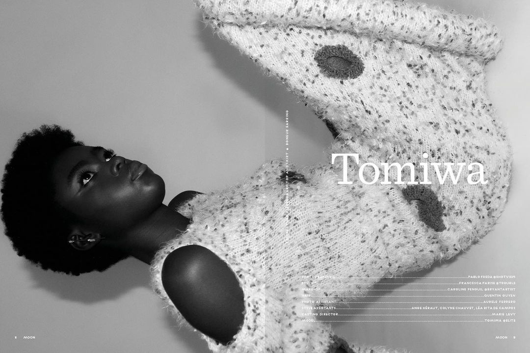 Portrait of a dark-skinned female model in a speckled sweater and black afro hairstyle looking up against a grey background in a studio.