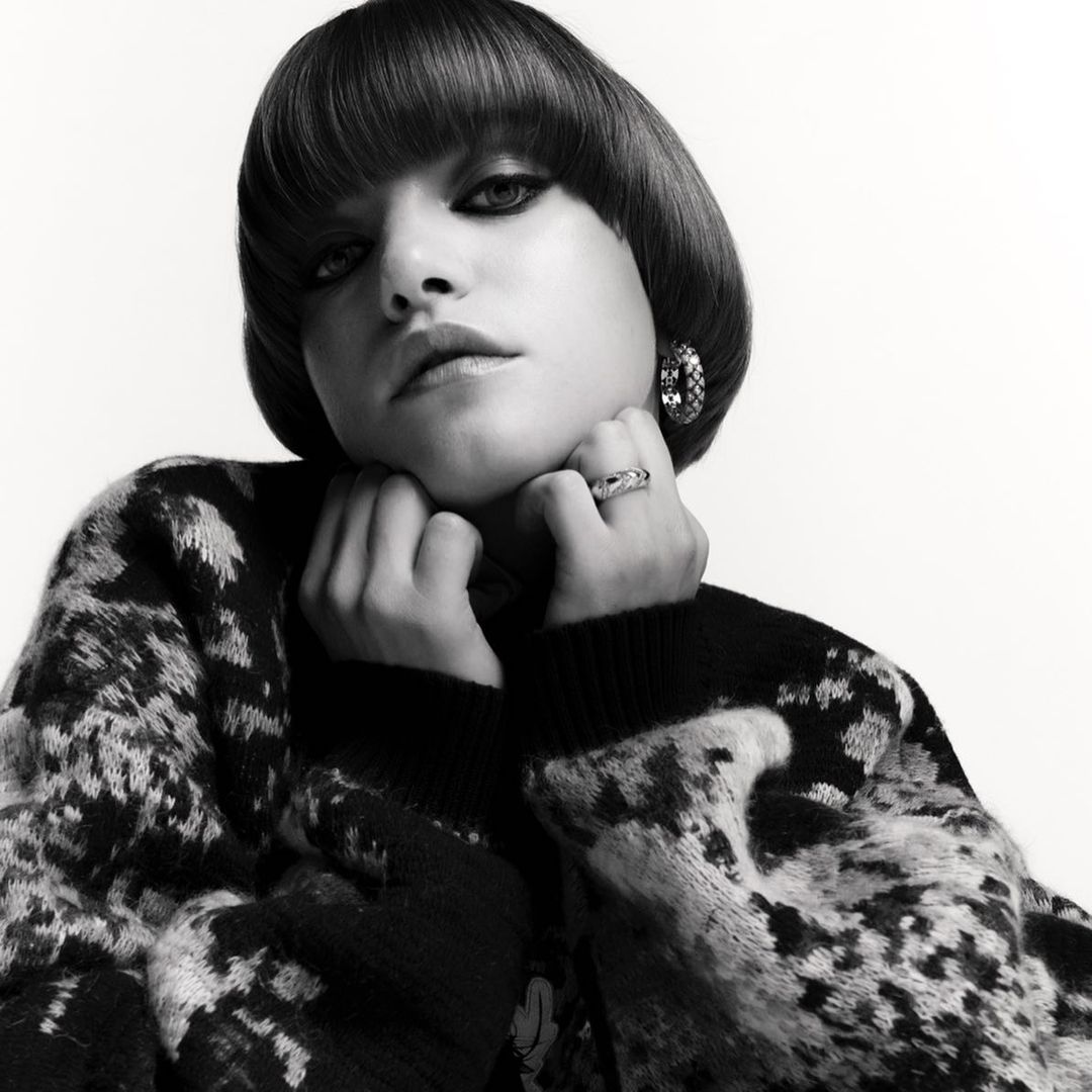 Portrait of a woman with a blunt bowl cut, wearing a patterned sweater and jewelry, posing against a plain white background in black and white.