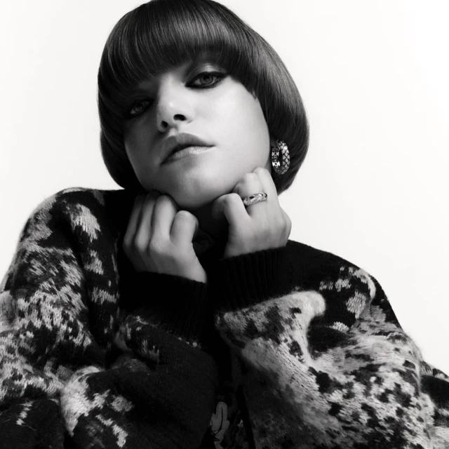 Portrait of a woman with a blunt bowl cut, wearing a patterned sweater and jewelry, posing against a plain white background in black and white.
