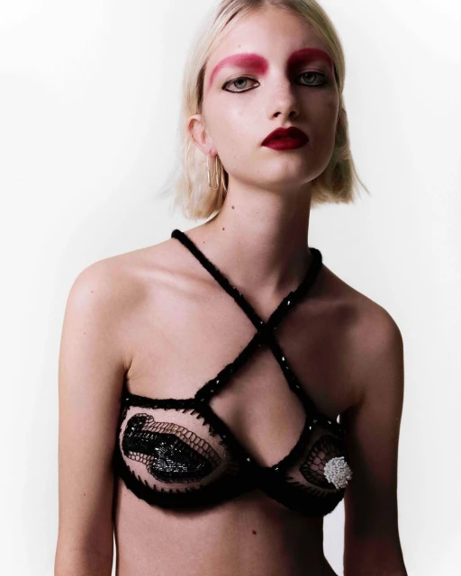 Woman with bleached blond hair and vivid red makeup posing in a crocheted halter top in front of a solid white backdrop.