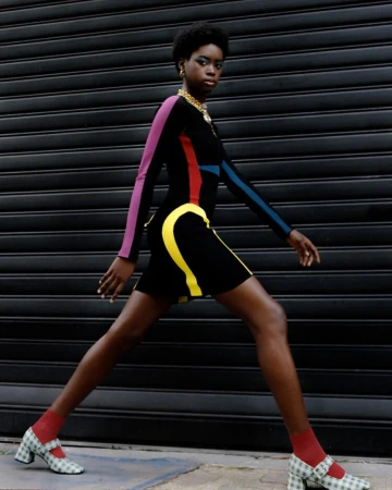 Model with short dark hair walking in a form-fitting color-blocked dress and checkered block heels against a dark shuttered storefront.