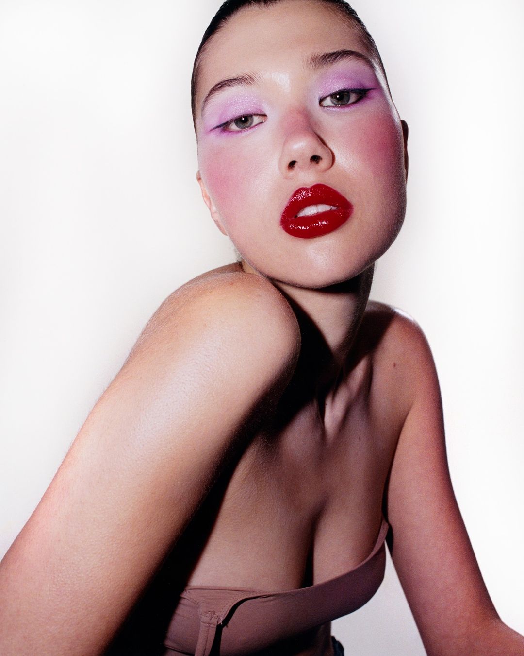Close-up of a beautiful young woman with slicked-back hair, wearing vibrant pink and red makeup, posing in a photo studio.