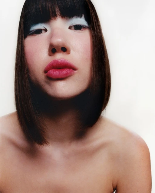Headshot of an East Asian woman with bob haircut, pale skin, bright pink lips, light blue eyeshadow, looking at the camera against white.