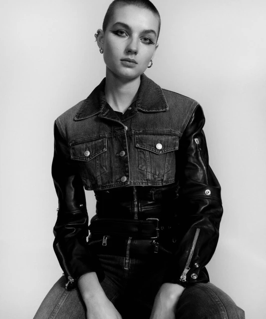 Monochromatic studio portrait of a woman with a shaved head wearing a denim and leather jacket and dark jeans against a white backdrop.