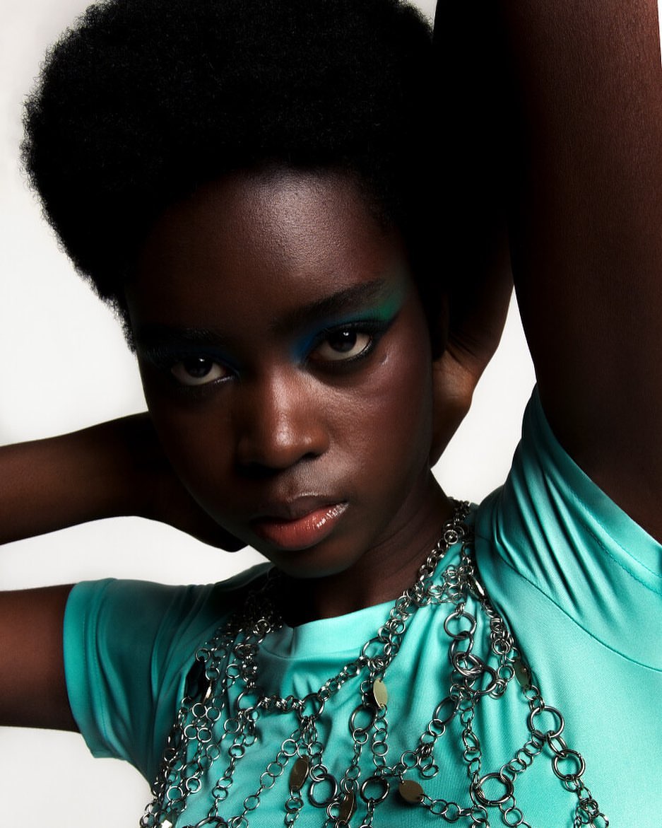A close-up studio portrait of a Black woman with green and blue eye makeup, wearing a teal shirt and a silver chain necklace.