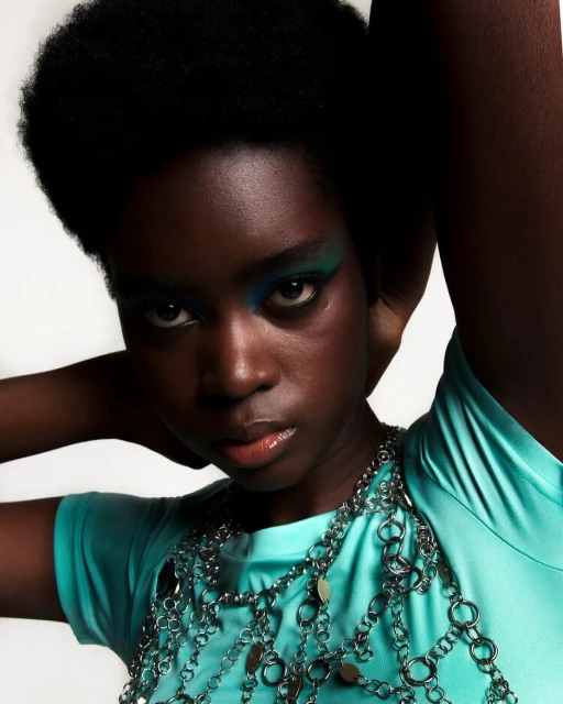 A close-up studio portrait of a Black woman with green and blue eye makeup, wearing a teal shirt and a silver chain necklace.