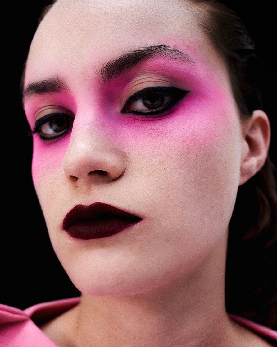 Close-up of a woman with pink eyeshadow, heavy black eyeliner, and dark red lipstick against a black background for beauty campaign.