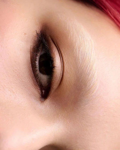 Close-up shot of an adult female's eye with brown iris and dark lashes, fair skin, and pink hair, possibly in a beauty or fashion context.