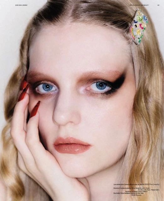 Close-up portrait of a young woman with long blonde hair and a hair accessory, wearing dramatic makeup and long red nails.