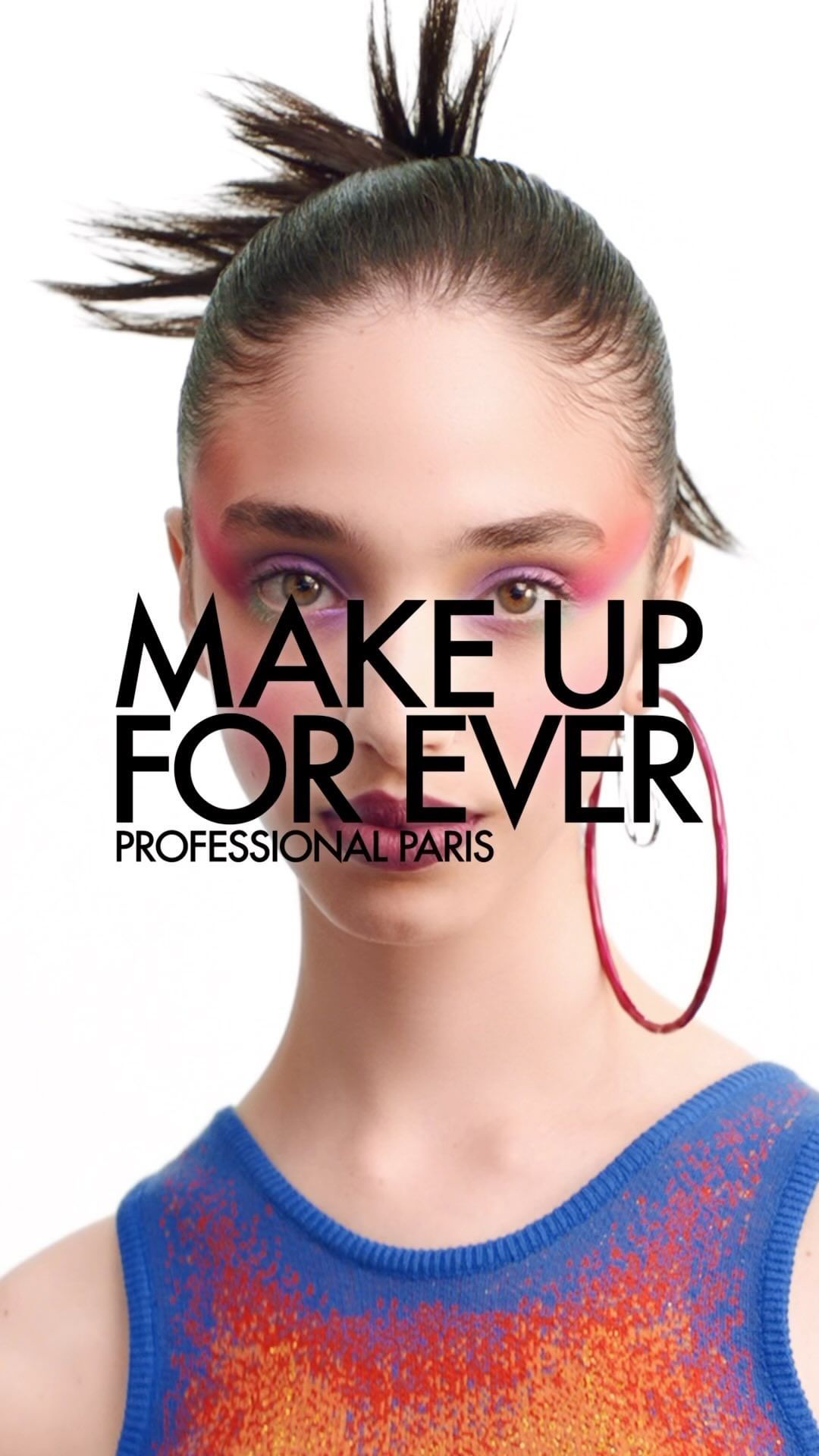 A woman is centered in a portrait wearing colorful eyeshadow and a blue and orange top. The background is white, and hoop earrings are visible.