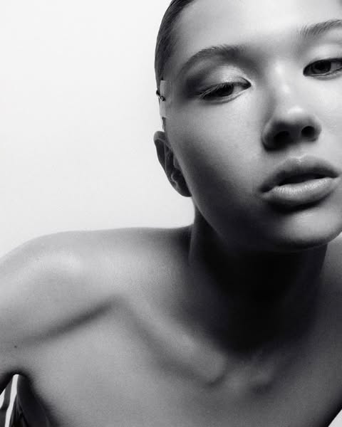Striking black and white portrait of a young adult female with a bare shoulder, gazing downward against a stark white backdrop in a photo studio.