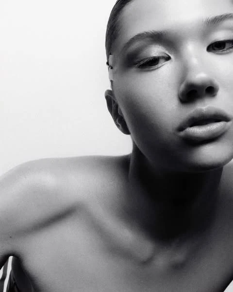 Striking black and white portrait of a young adult female with a bare shoulder, gazing downward against a stark white backdrop in a photo studio.