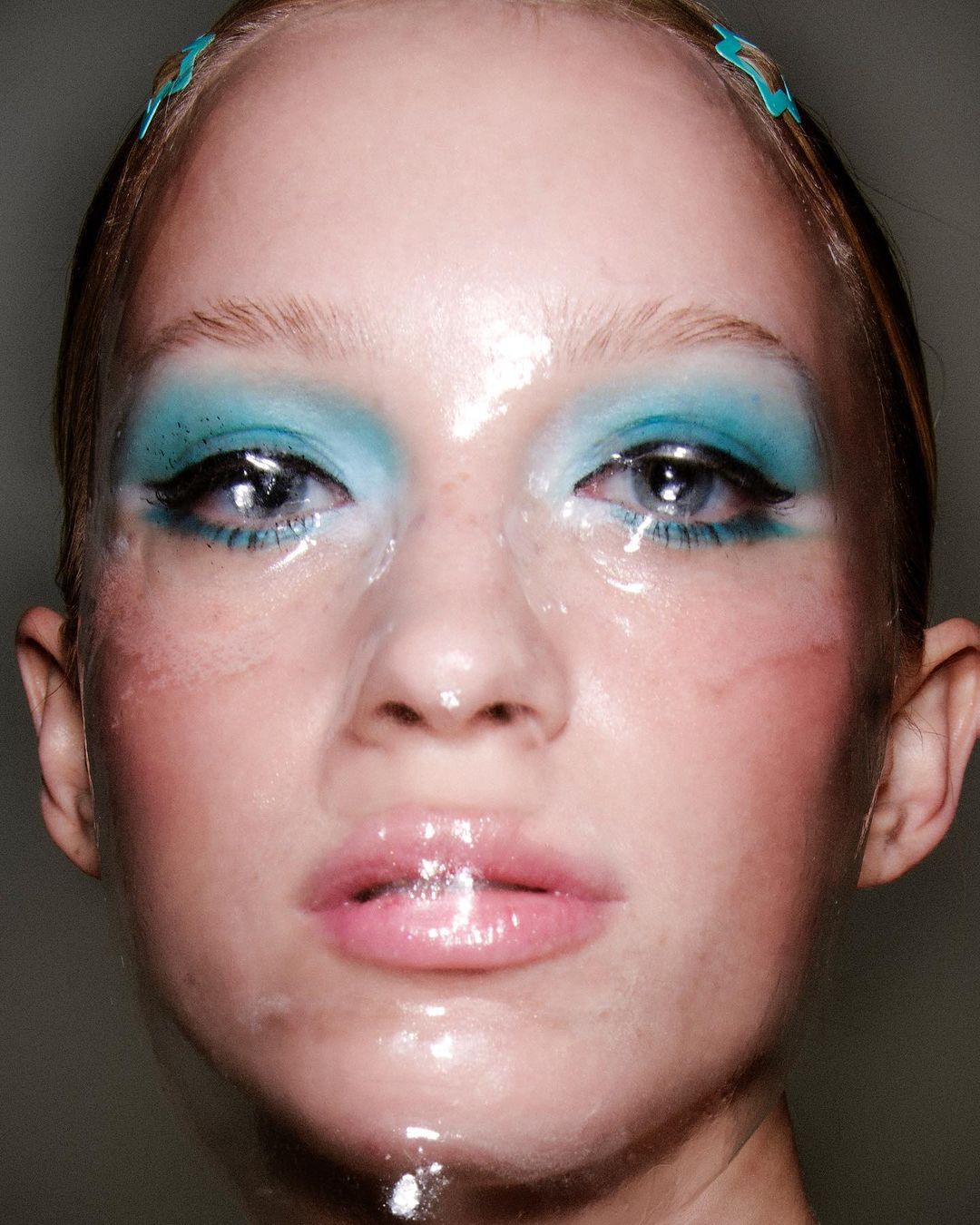 Close-up portrait of a woman with cyan eyeshadow, light pink lips, and a shiny film covering her face and head.
