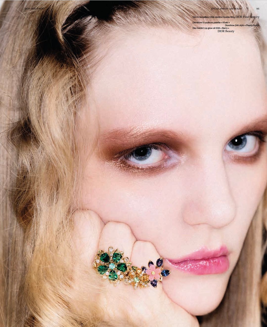 Close-up of a woman with fair skin, makeup featuring brown eyeshadow and pink lipstick, and a large floral ring on her finger.