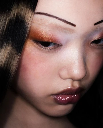 Close-up portrait of a model with thin drawn-on eyebrows, warm shimmering eyeshadow, and glossy dark lips by Pablo Freda.