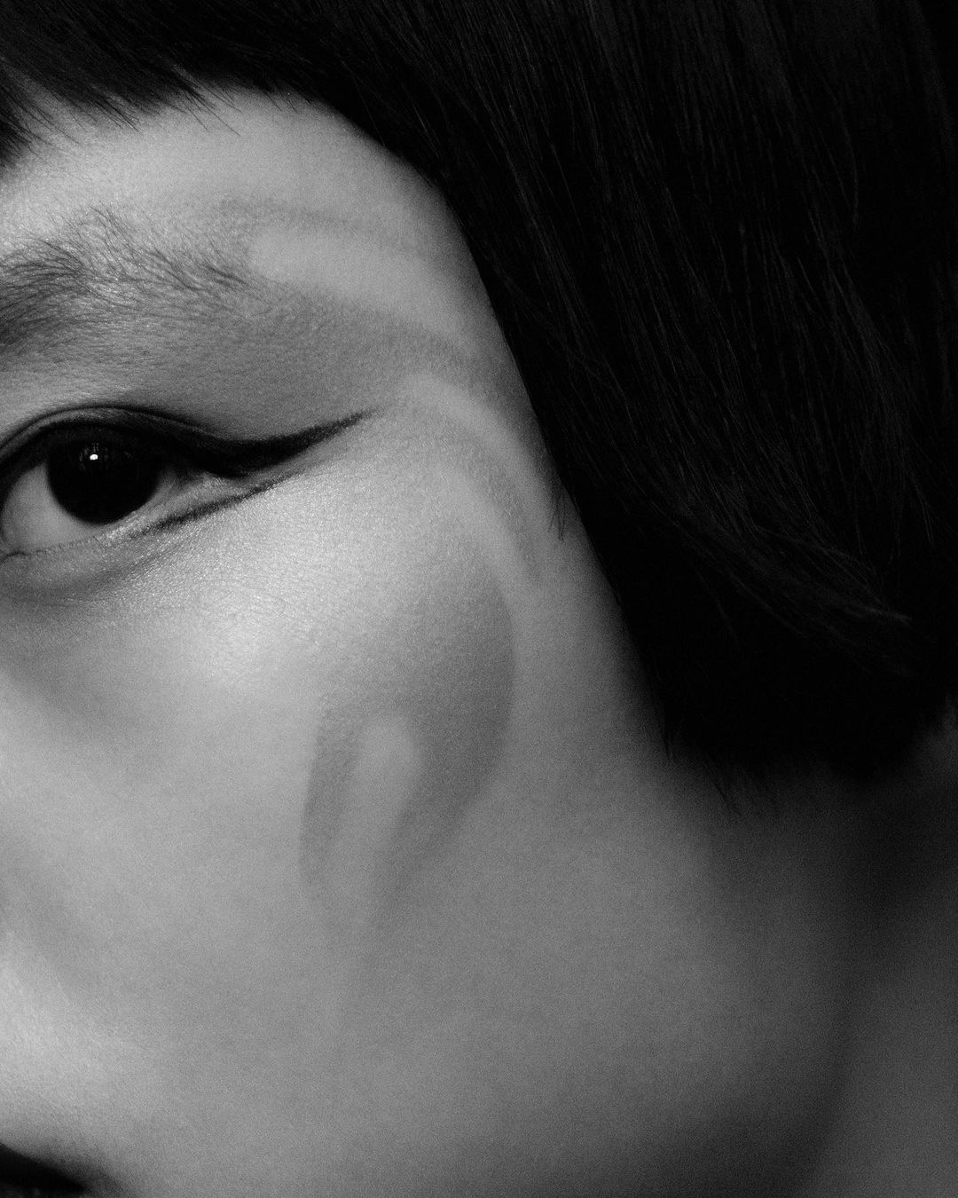 Monochrome close-up of a woman's face focusing on the eye with dramatic eyeliner, with short black hair and subtle cheek highlighting.