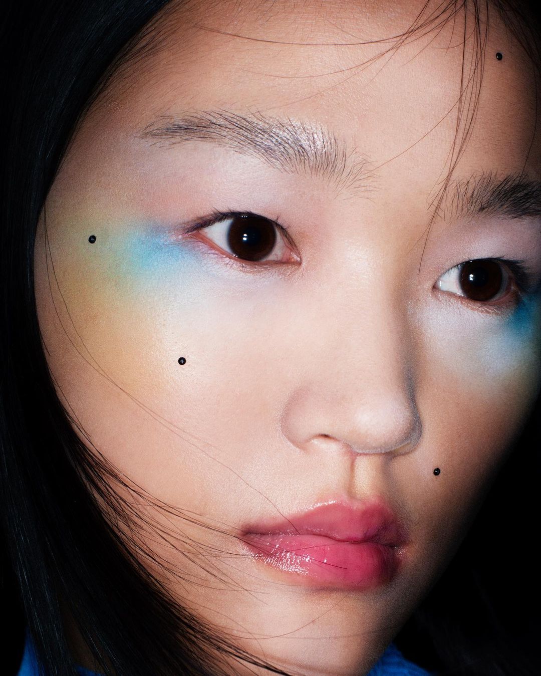 Close-up portrait of an Asian woman with vibrant blue and yellow eyeshadow, glossy pink lips, and dark hair against a dark backdrop.