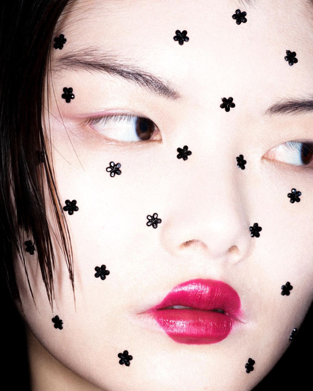 Close-up portrait of an Asian female model wearing floral facial decorations and bold fuchsia lipstick against a dark background.