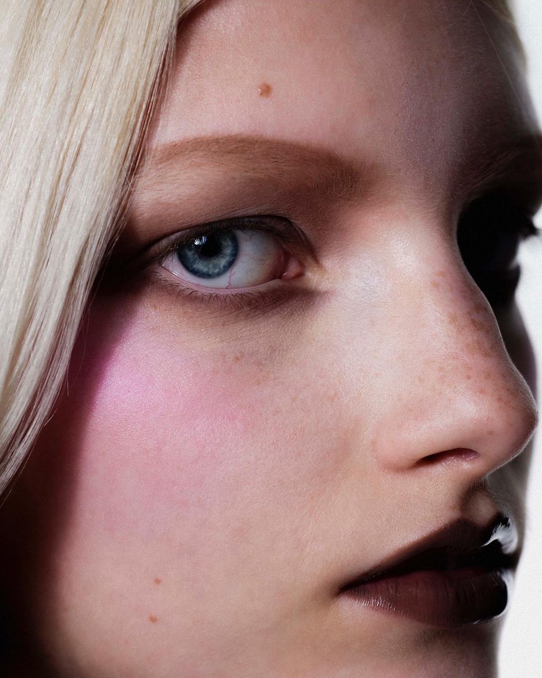 Close-up beauty shot of a fair-skinned adult woman with blonde hair and blue eyes, featuring a pink cheek and dark lip makeup.