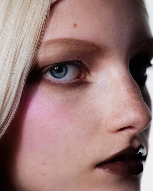 Close-up beauty shot of a fair-skinned adult woman with blonde hair and blue eyes, featuring a pink cheek and dark lip makeup.