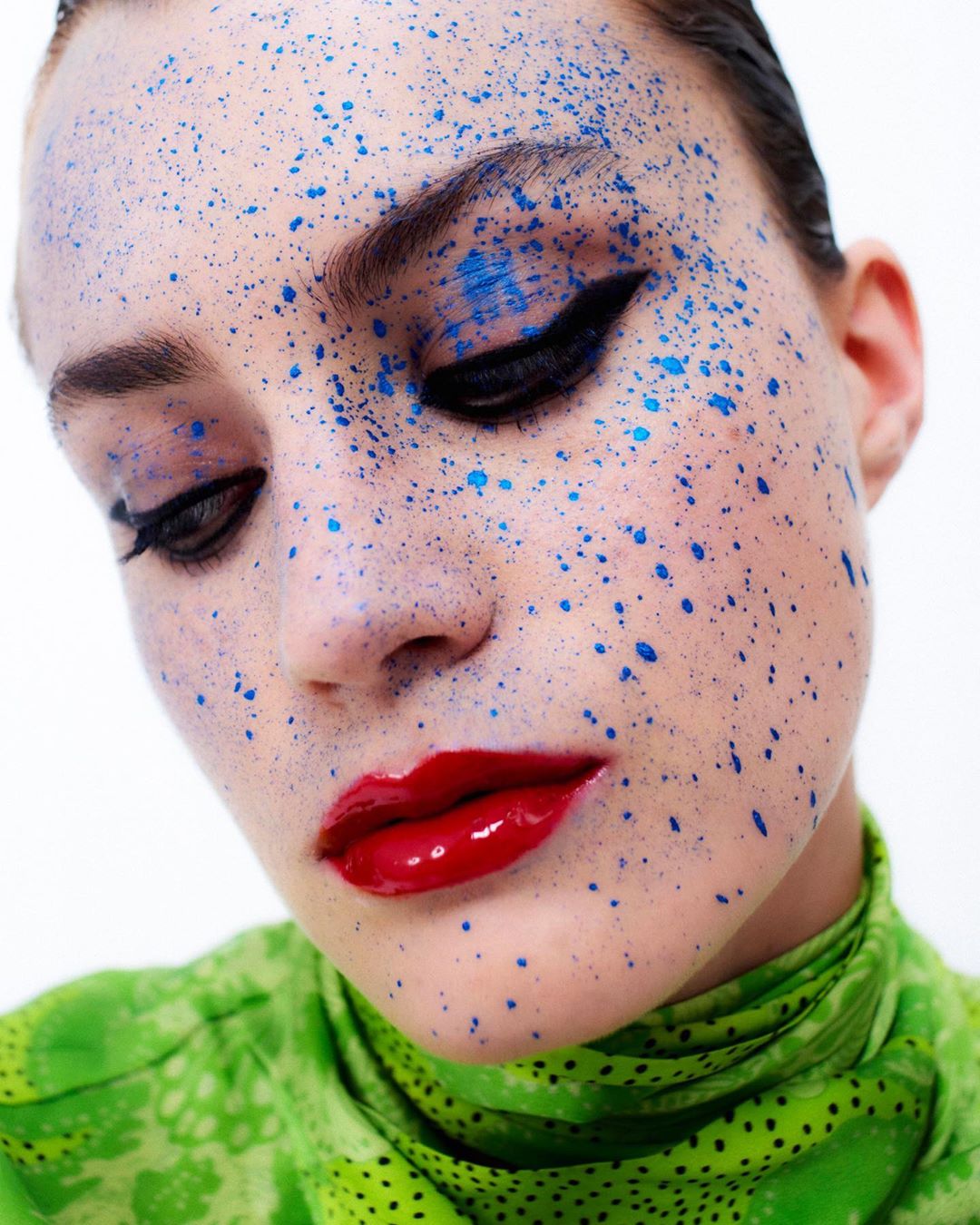 Close-up portrait of a woman with blue paint splatters on her face, glossy red lips, wearing a green patterned top against a white backdrop.
