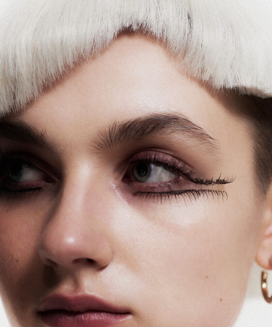 Close-up portrait of a young woman with neutral expression, wearing a white furry hat, with dramatic black eyeliner and a gold hoop earring.